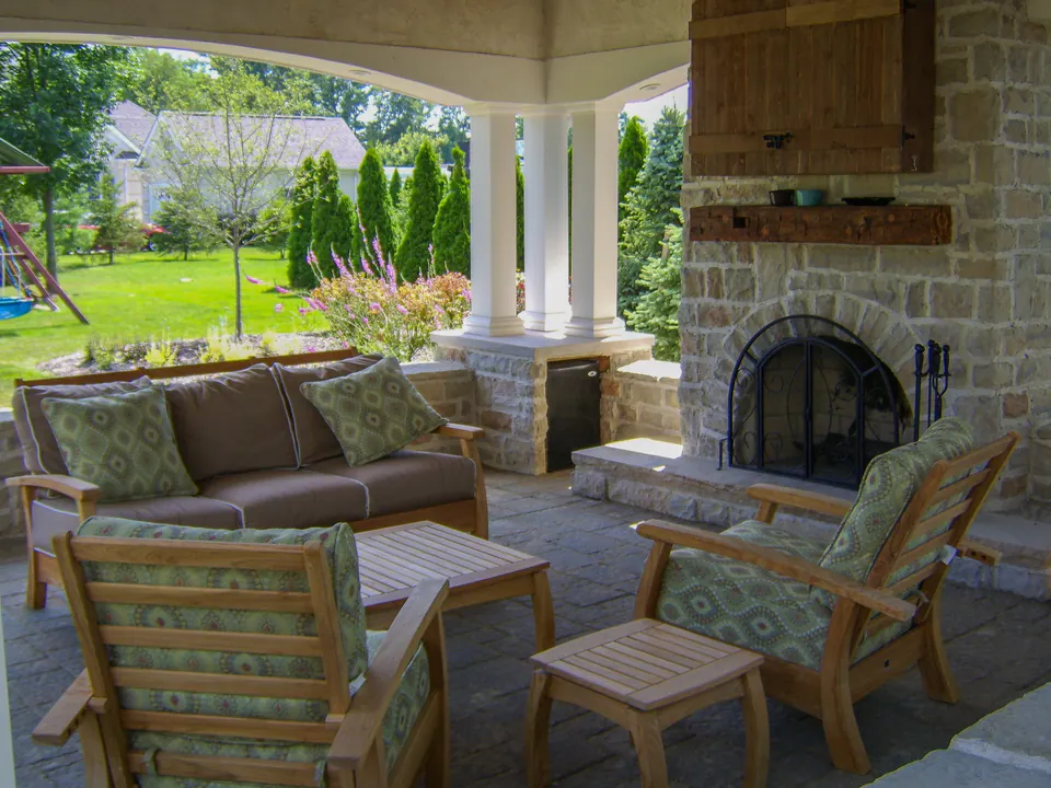Covered seating area with stone fireplace, paver floor, sofa, chairs, columns, and garden view.